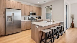 Una cocina moderna con armarios de madera, electrodomésticos de acero inoxidable, una isla central con taburetes altos y un ventanal que proporciona luz natural.