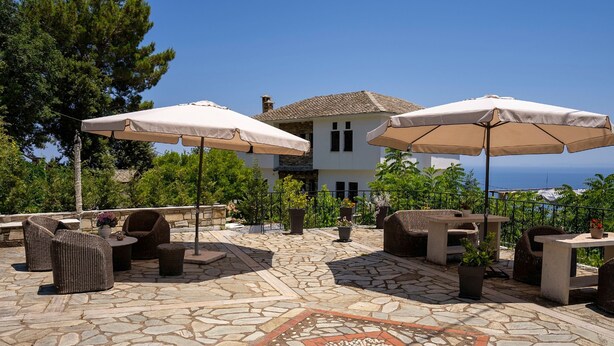 A patio area with stone flooring, outdoor furniture, and umbrellas, overlooking a house and greenery. - Paradeisos Double Room With Balcony 2 (Zagora-Mouresi)