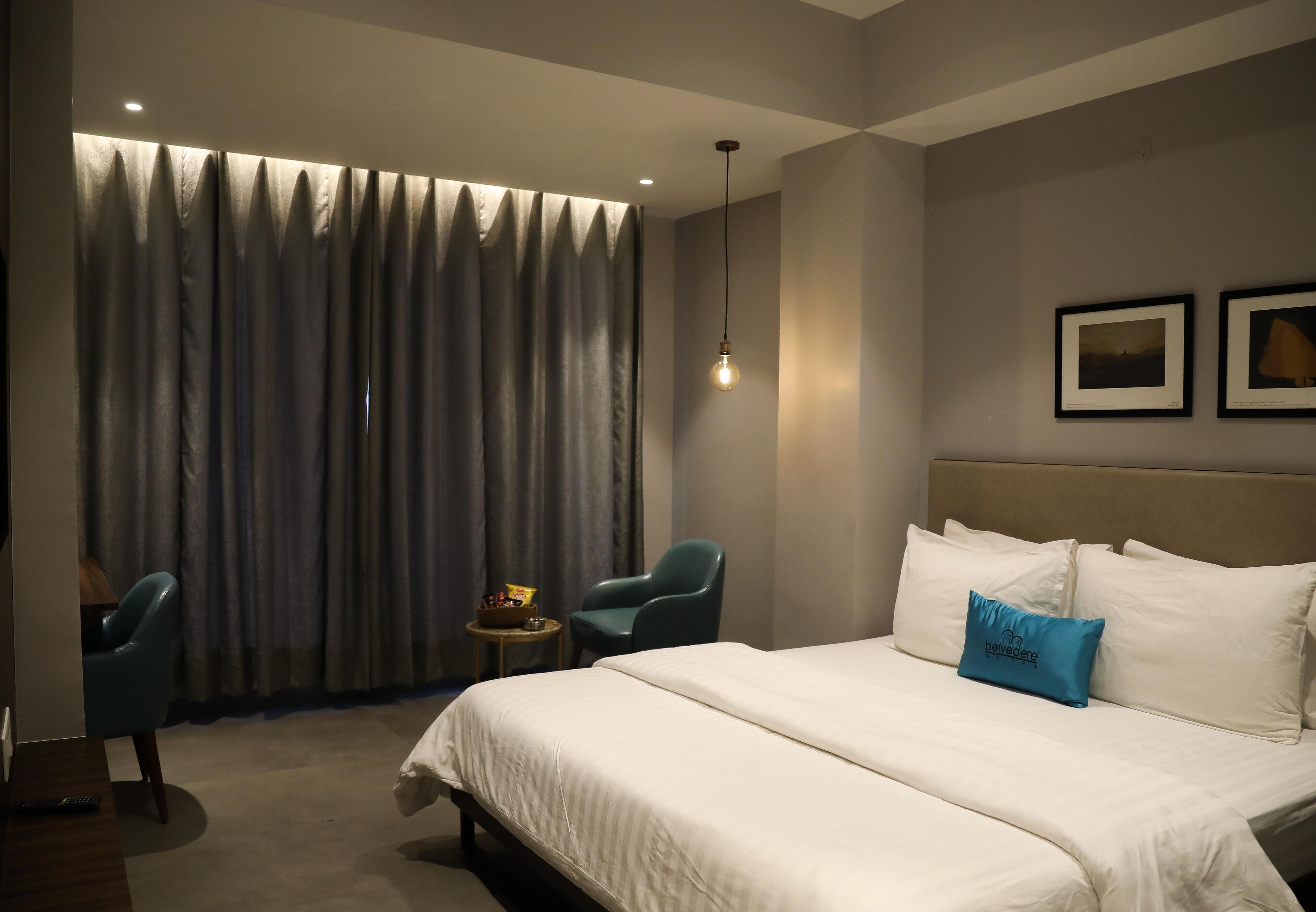 Une chambre d’hôtel avec un grand lit, deux chaises, une petite table et un rideau agrémenté d’un luminaire.