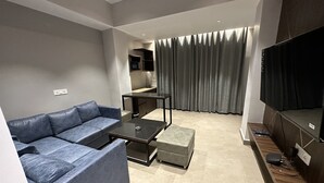 En moderne stue med en grå sofa, et sofabord, et fjernsyn og et køkken i baggrunden.