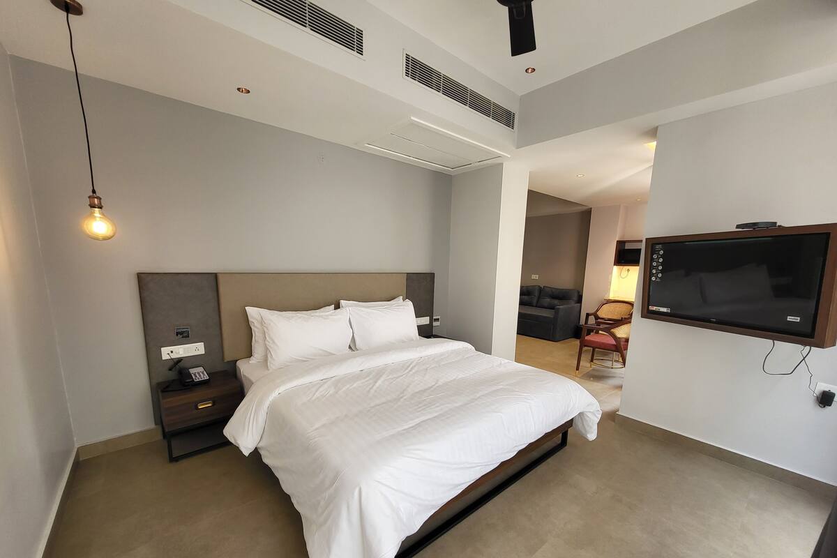 A modern hotel room with a large bed, a bedside table with a phone, a TV mounted on the wall, and a ceiling vent.