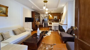 A modern living room with a sofa, a coffee table, and a TV. There is a kitchen area with wooden cabinets and a refrigerator. - Harmony Platanidia Pelion (Volos)