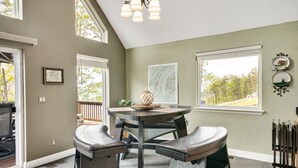 A dining area with a round table, two cushioned benches, a chandelier, and a view of the outdoors through large windows.