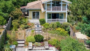Una casa de dos pisos con balcón, rodeada de abundante vegetación y una terraza de madera.