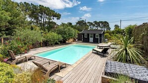 Una piscina rectangular con una terraza de madera, sillas de descanso y una pequeña casa al fondo.