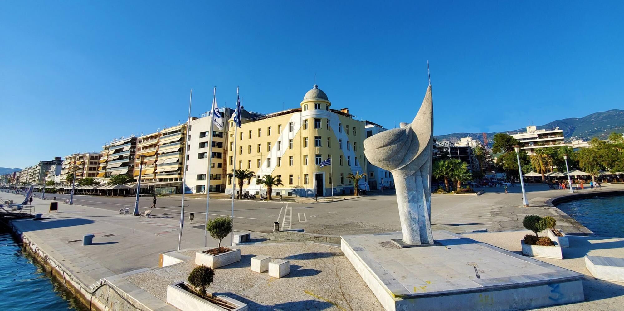 A waterfront promenade with a large sculpture, a yellow building with multiple floors, and a clear blue sky.