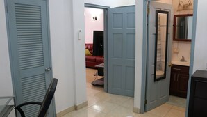 Un couloir avec une porte fermée sur la gauche, une porte ouverte donnant sur un salon avec un canapé rouge, et une salle de bain où l’on peut voir un miroir et un lavabo.