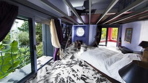Ein Schlafzimmer mit einem großen Bett, einem Fenster mit Blick auf Grünflächen, einem runden Fenster und einer Treppe zu einer oberen Ebene.