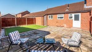 A backyard with a patio, two striped lounge chairs, a table with a bottle and wine glasses, and a well-maintained lawn.
