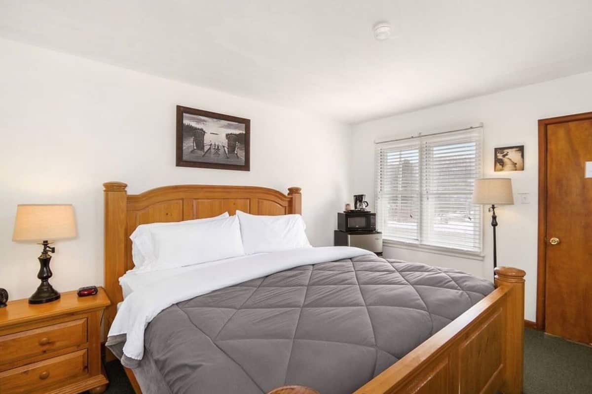 A neatly made bed with a grey quilt, a wooden headboard, a bedside table with a lamp, a small picture frame, and a window with blinds.