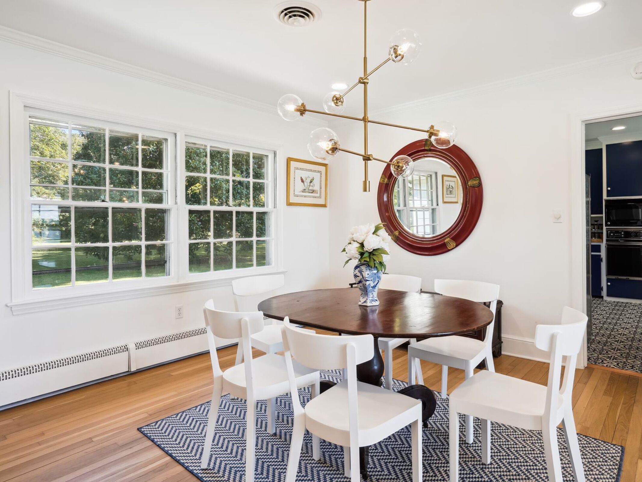 A dining room with a round table, white chairs, a vase with flowers, a chandelier, and a large window with multiple panes.