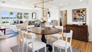 A spacious living room with a round dining table surrounded by white chairs, a large window with a view of a lake, and a modern chandelier. - Minutes from Downtown Easton and St. Michaels | Pool | Private Dock (Easton)