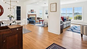A spacious living room with hardwood floors, a fireplace, a dining area with a wooden table and chairs, and a view of the outdoors through large windows. - Minutes from Downtown Easton and St. Michaels | Pool | Private Dock (Easton)