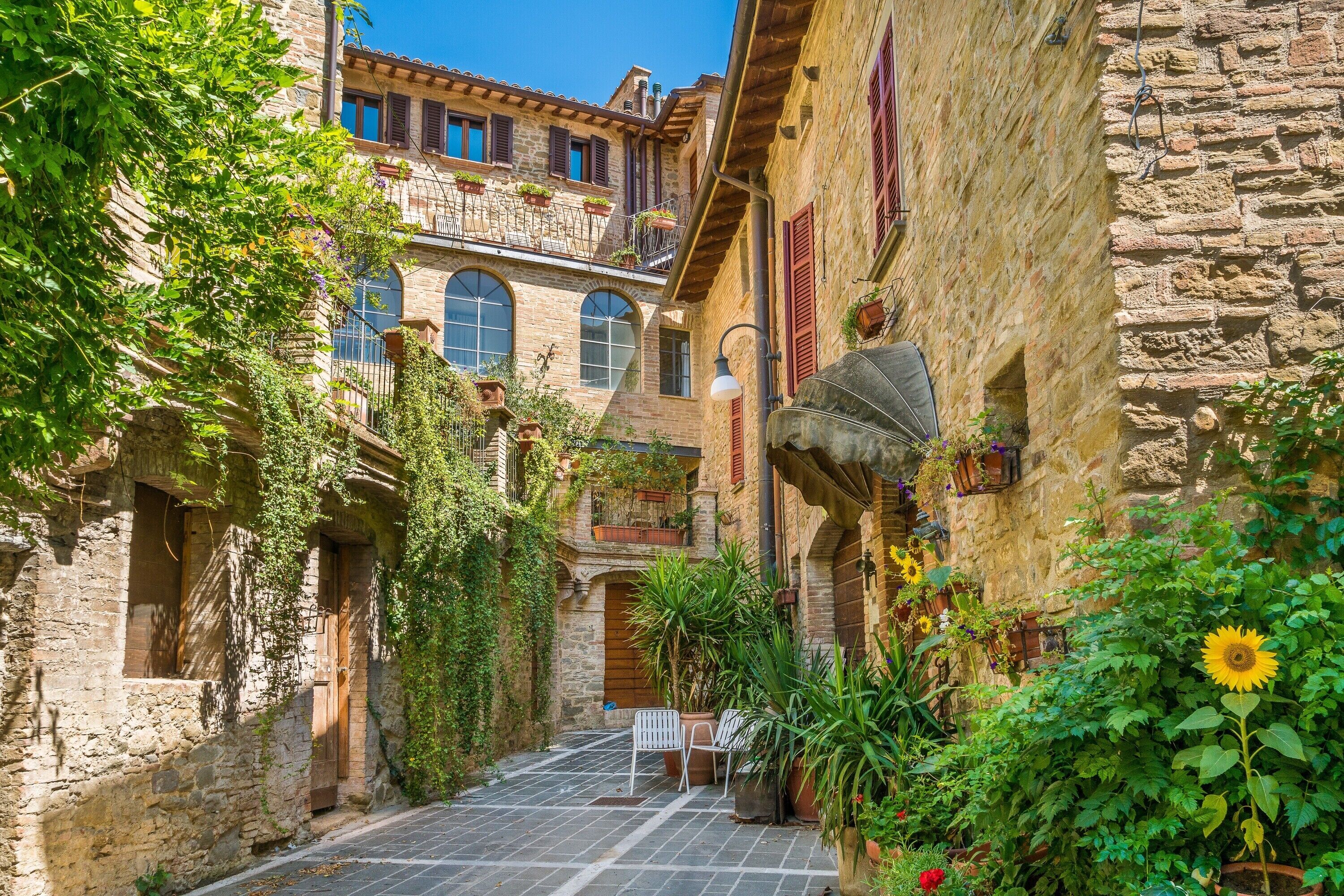A narrow alley with stone buildings, a small table and chairs, and potted plants.