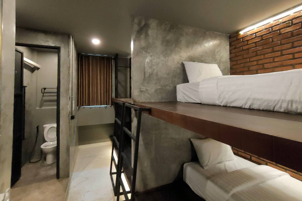 Ein kompaktes Hotelzimmer mit Etagenbett, einem kleinen Badezimmer inklusive Toilette und einem Fenster mit Jalousien.