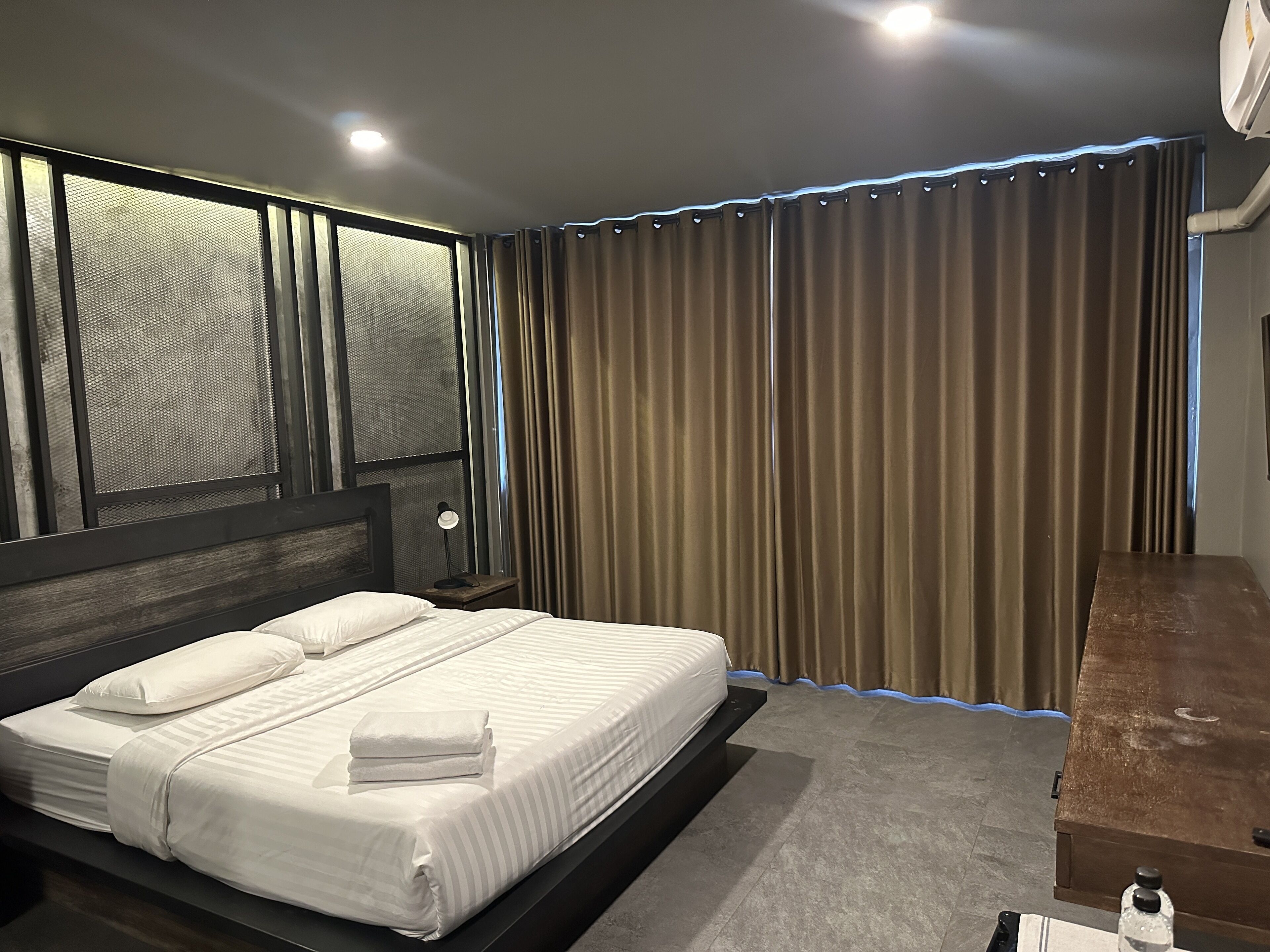 Une chambre moderne avec un grand lit, une tête de lit au motif texturé, une table de chevet avec une lampe et une fenêtre donnant sur l’extérieur.