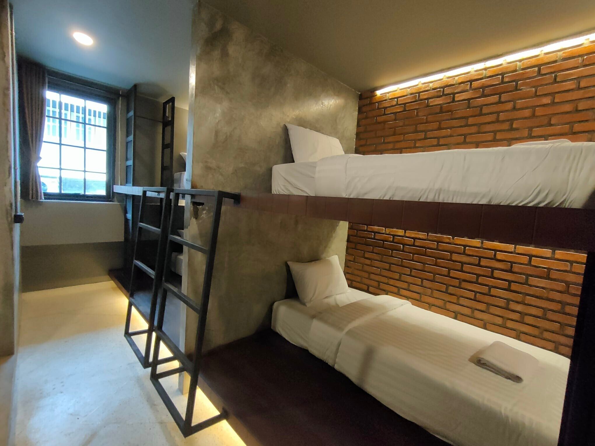 A compact room with bunk beds, a ladder, a window with curtains, and a brick-patterned wall.