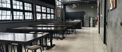A spacious room with long tables and stools, large windows, a kitchen area, and a high ceiling with exposed beams.