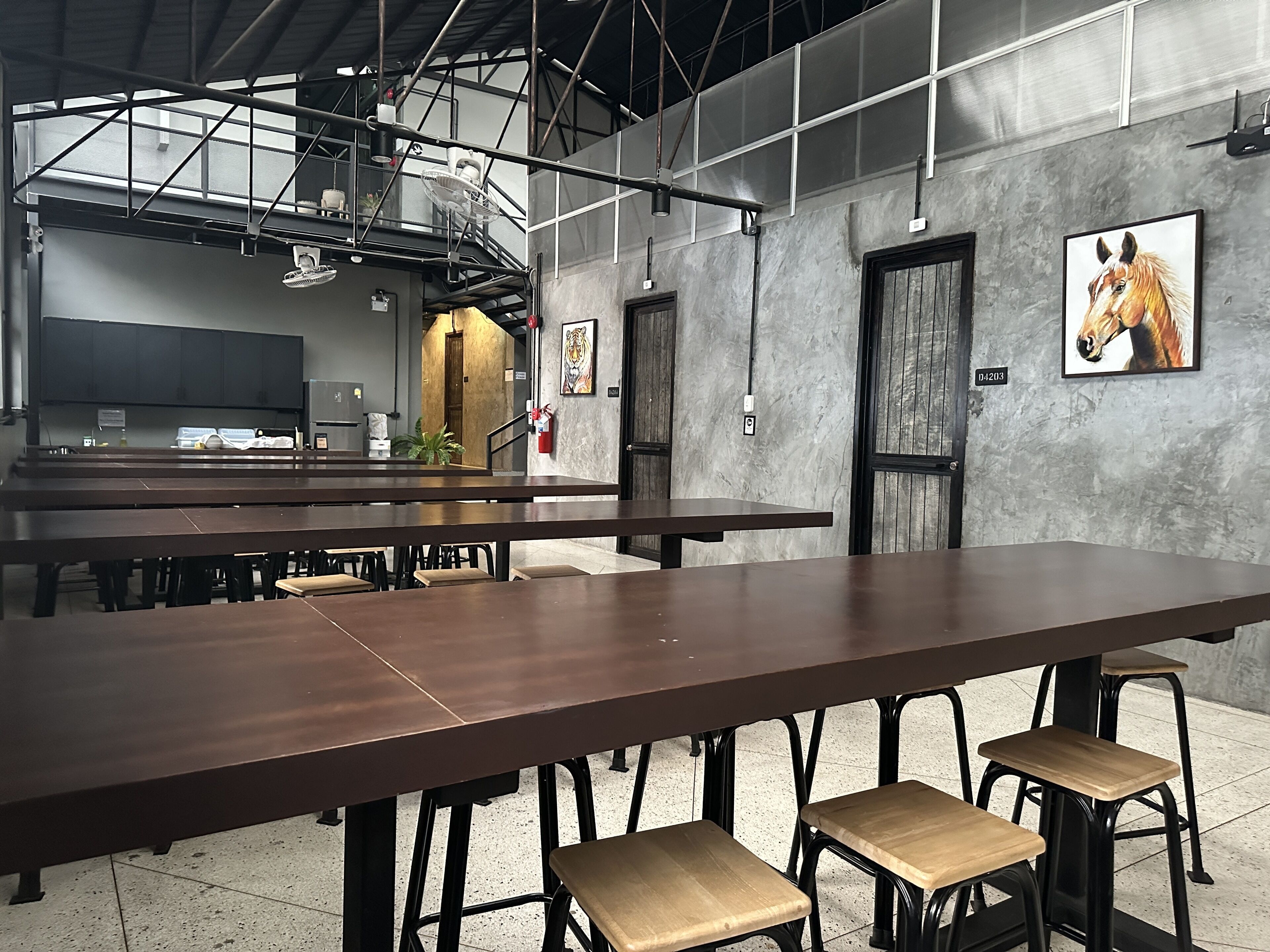 Un grand espace intérieur avec une longue table et plusieurs chaises, un extincteur et un tableau représentant un cheval accroché au mur.