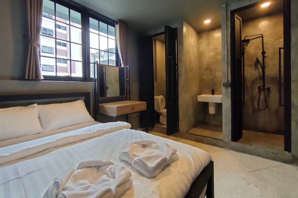 Ein Hotelzimmer mit Bett, Nachttisch, Spiegel, Fenster mit Vorhängen sowie einem Badezimmer mit Waschbecken und Dusche.