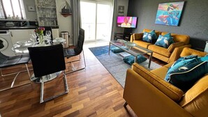 Uma sala de estar moderna com um sofá amarelo, uma mesa de centro de vidro e uma TV. Há uma área de jantar com uma mesa posta para dois e uma cozinha visível ao fundo.