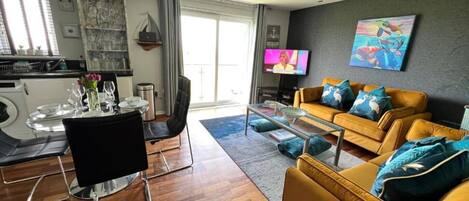 A modern living room with a yellow sofa, a glass coffee table, and a TV. There is a dining area with a table set for two and a kitchen visible in the background.