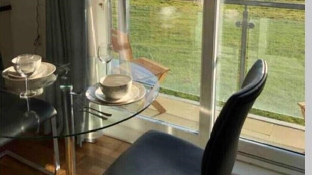 Uma área de refeições moderna com mesa e cadeiras de vidro, vista para o mar e paisagem gramada.