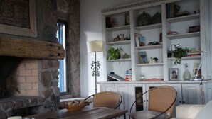 A cozy room with a stone fireplace, wooden table, wicker chairs, and a white bookshelf filled with books and decorative items.