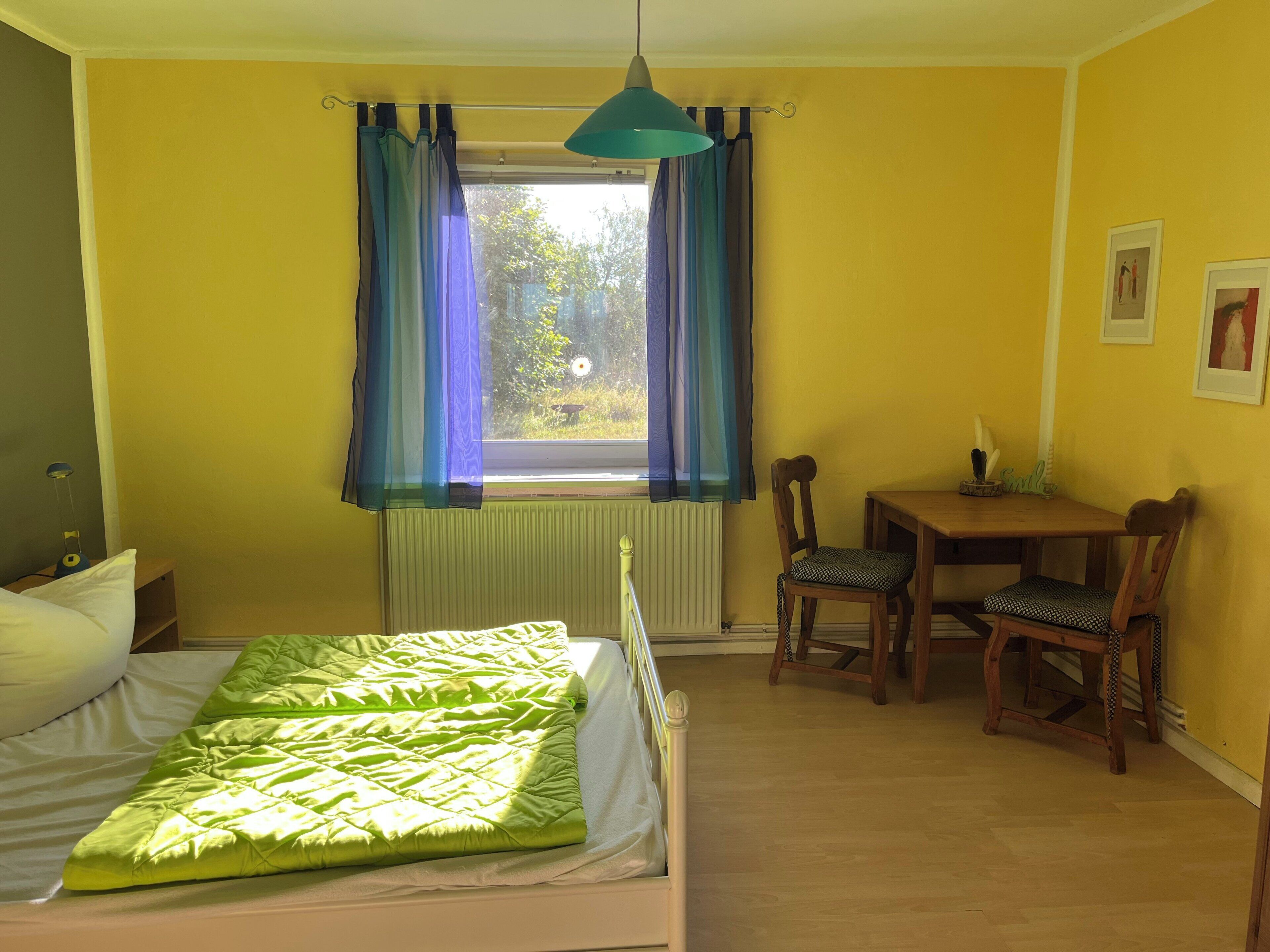 Una habitación con cama, una mesa, dos sillas, un ventanal con cortinas y un radiador.