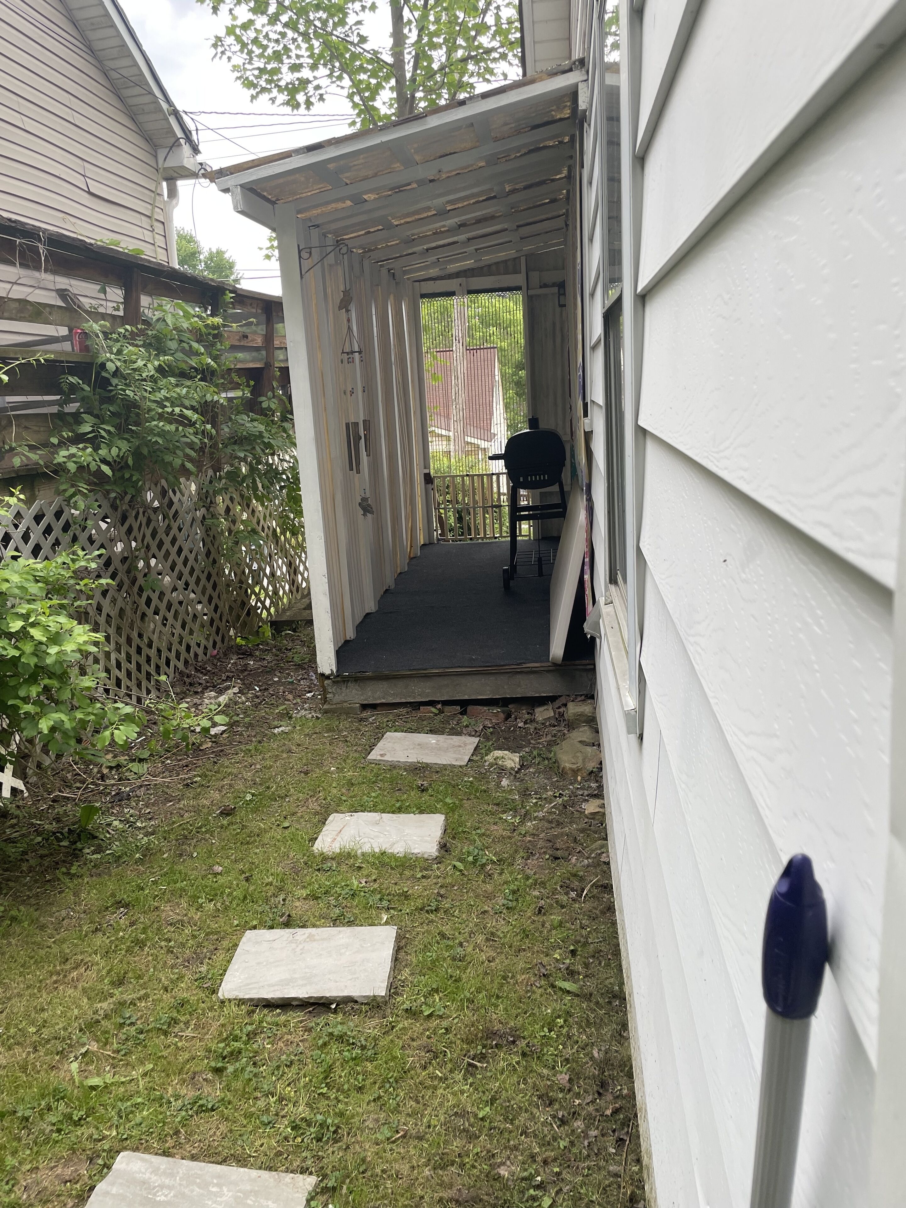 Un espace extérieur abrité, doté d’un chemin en béton menant à une porte, et entouré d’une clôture en bois et de verdure.