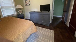 Un dormitorio con cama, cómoda, televisor, lámpara y una obra de arte en la pared.