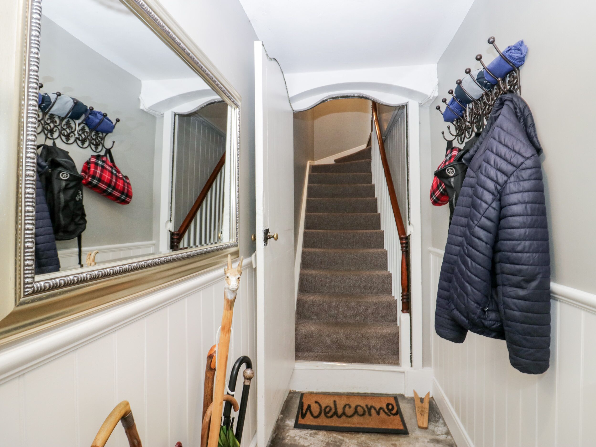 A hallway with a mirror, a coat rack with jackets, a wooden cane, and a welcome mat.