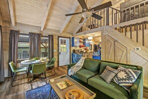 Un salon chaleureux avec un canapé vert, une table à manger entourée de chaises et un escalier menant à l’étage supérieur.