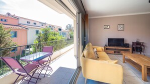 A balcony with a glass table and chairs, a yellow sofa, a wooden coffee table, and a TV on a wooden stand.
