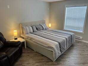A bedroom with a bed, two bedside lamps, a leather chair, a window with blinds, and a wall-mounted heater. - King Suite with Fireplace
Upper Floor Suite (Three Hills)
