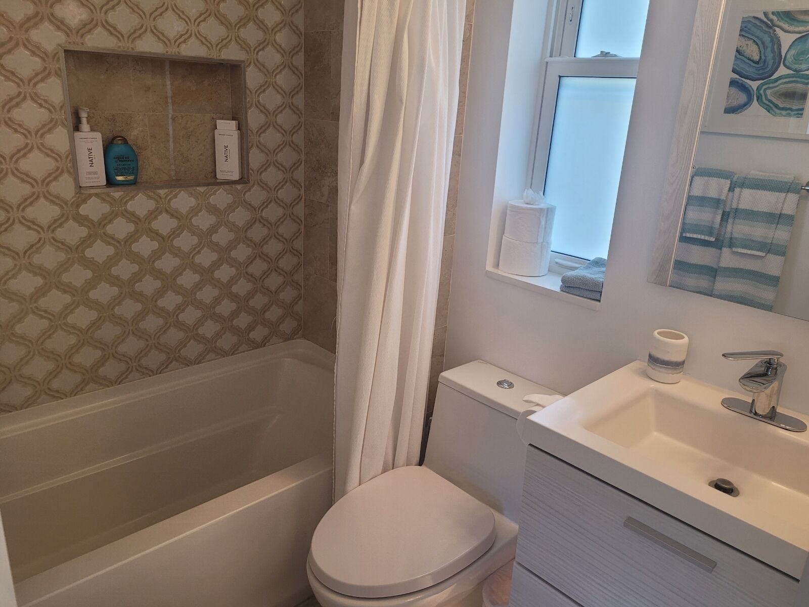 Ein Badezimmer mit Badewanne, Toilette und Waschbecken. Ein Ablagefach mit Pflegeartikeln und ein Handtuchhalter sind vorhanden.