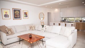Ein modernes Wohnzimmer mit einem weißen Ecksofa, einem Holztisch und einer Küchenzeile mit weißen Schränken und einem großen Fenster.