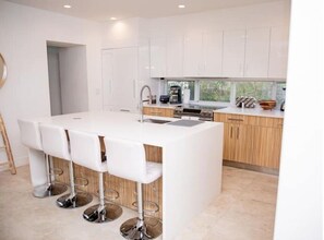 A modern kitchen with a central island, bar stools, wooden cabinets, and a window providing natural light. - 5 star Beachside Home Next to Everything! Patio/Wifi/TVs (Lauderdale-by-the-Sea)