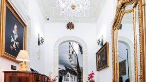 Un vaste hall d’entrée orné d’un lustre, de miroirs décoratifs et d’un paillasson noir sur lequel est inscrit le mot « NOBILIS ».