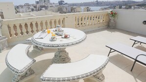 A round table with a white and black patterned tablecloth, two white cushioned benches, and a potted plant with red flowers. - Deluxe Apartment in Nobilis Guesthouse Apt 4 (Sliema)