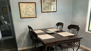 Un coin repas avec une table et des chaises en bois, des cartes encadrées au mur et une fenêtre donnant sur un paysage.