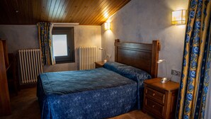 A bedroom with a wooden ceiling, a bed with a blue bedspread, a nightstand with a lamp, a window with curtains, and a radiator. - Hostal Cal Franciscó (Gosol)