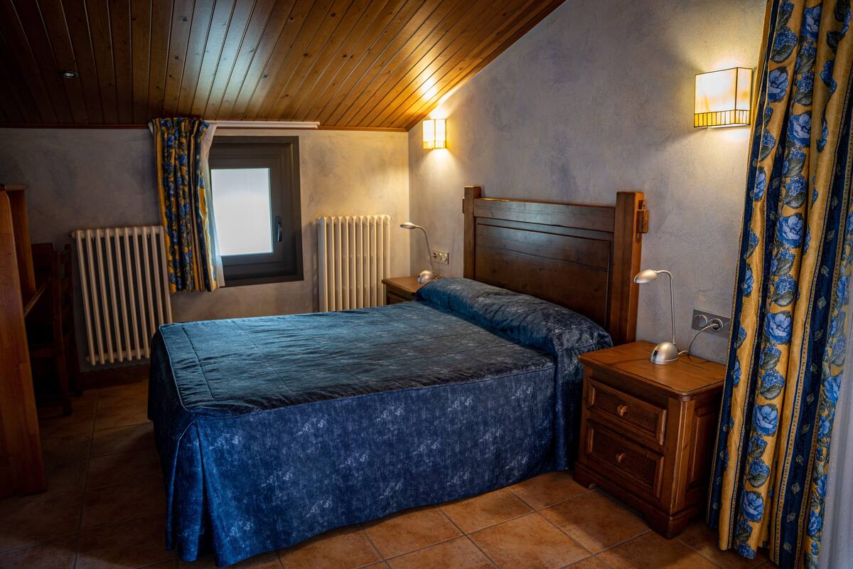 A bedroom with a wooden ceiling, a bed with a blue bedspread, a nightstand with a lamp, a window with curtains, and a radiator.