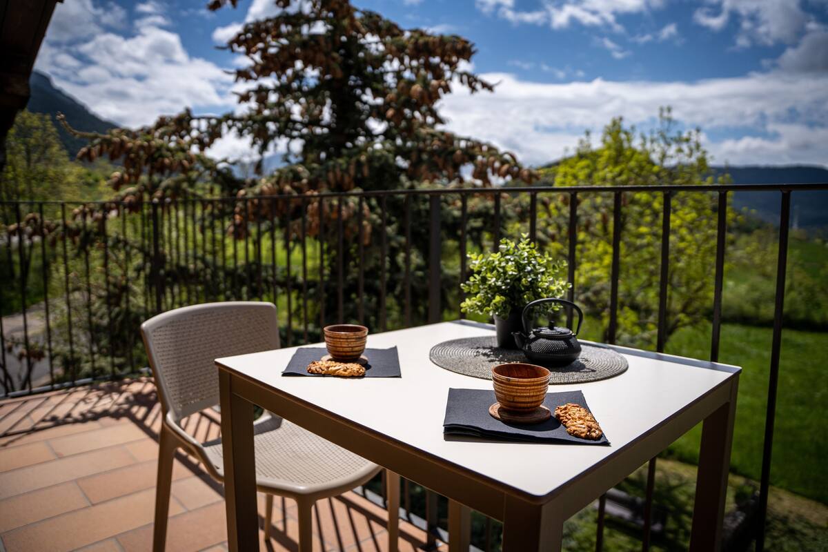 A table with two bowls, a teapot, and a plant on a balcony with a view of mountains and greenery.