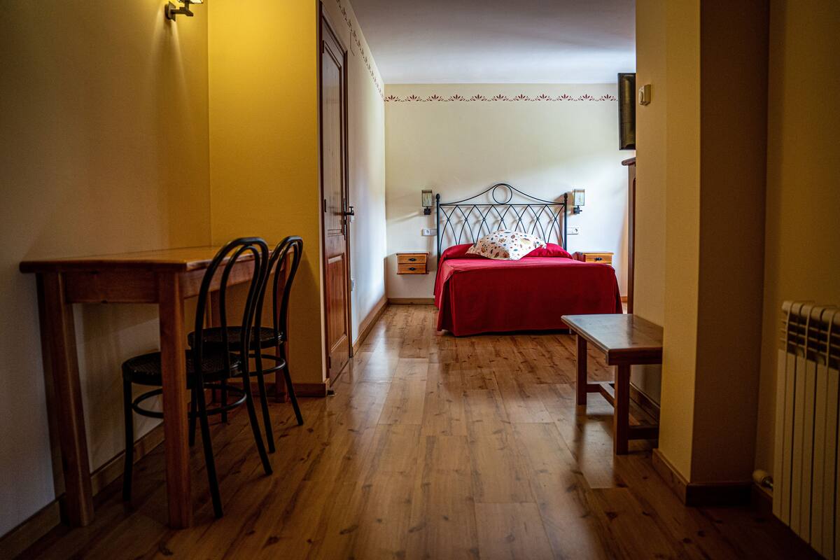 A room with a wooden floor, a bed with a red bedspread, a desk with a chair, and a nightstand.