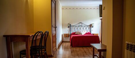 Una habitación con piso de madera, una cama con colcha roja, un escritorio con silla y una mesita de noche.