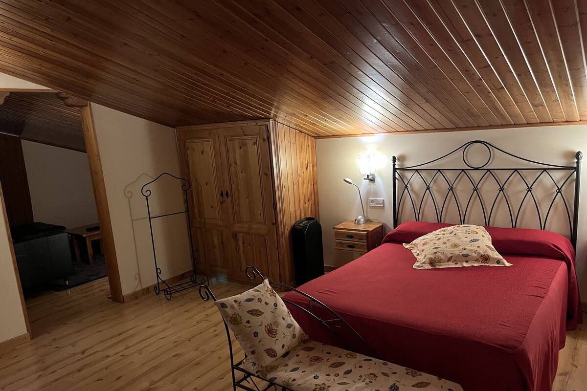 A bedroom with a wooden ceiling, a bed with red bedding, a bedside table with a lamp, and a chair with floral upholstery.