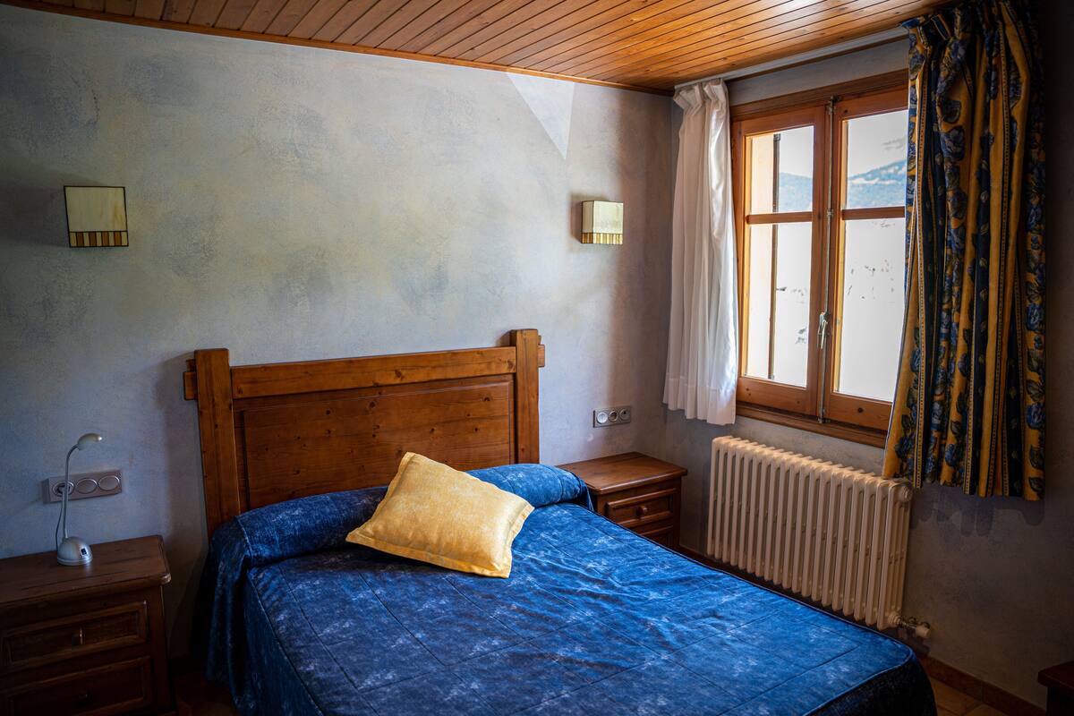 A bedroom with a wooden headboard, a blue bedspread, a single yellow pillow, bedside lamps, a nightstand, and a window with curtains.
