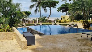Piscina com espreguiçadeiras e palmeiras, cercada por um muro de pedra e vista para a praia.