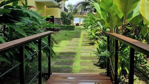 Uma passagem leva a um edifício com fachada amarela, rodeado por vegetação exuberante e palmeiras.